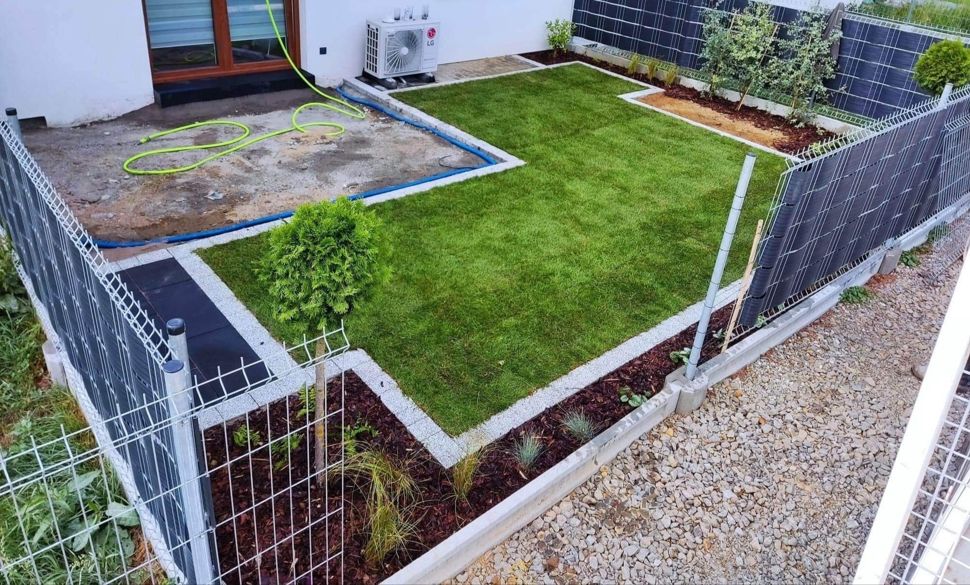 Świeżo założony trawnik z obrzeżami z jasnego grysu, widok z góry na ogród z siatką ogrodzeniową i roślinami ozdobnymi, obok domu z klimatyzatorem.