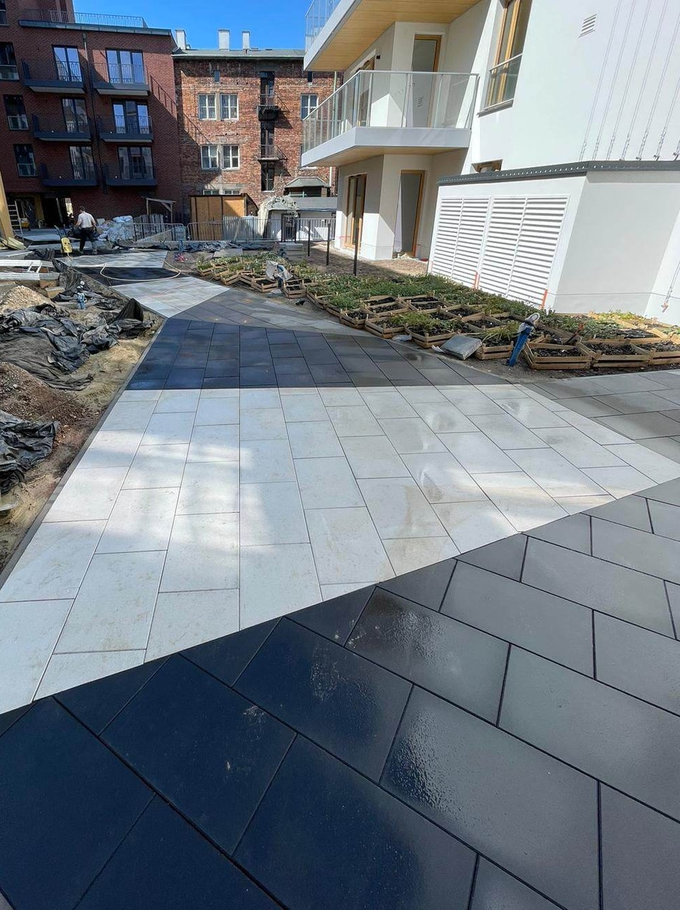 Nowoczesna nawierzchnia z jasnych i ciemnych płyt betonowych ułożonych we wzór, z widocznymi sadzonkami w drewnianych skrzynkach w tle nowego budynku mieszkalnego.