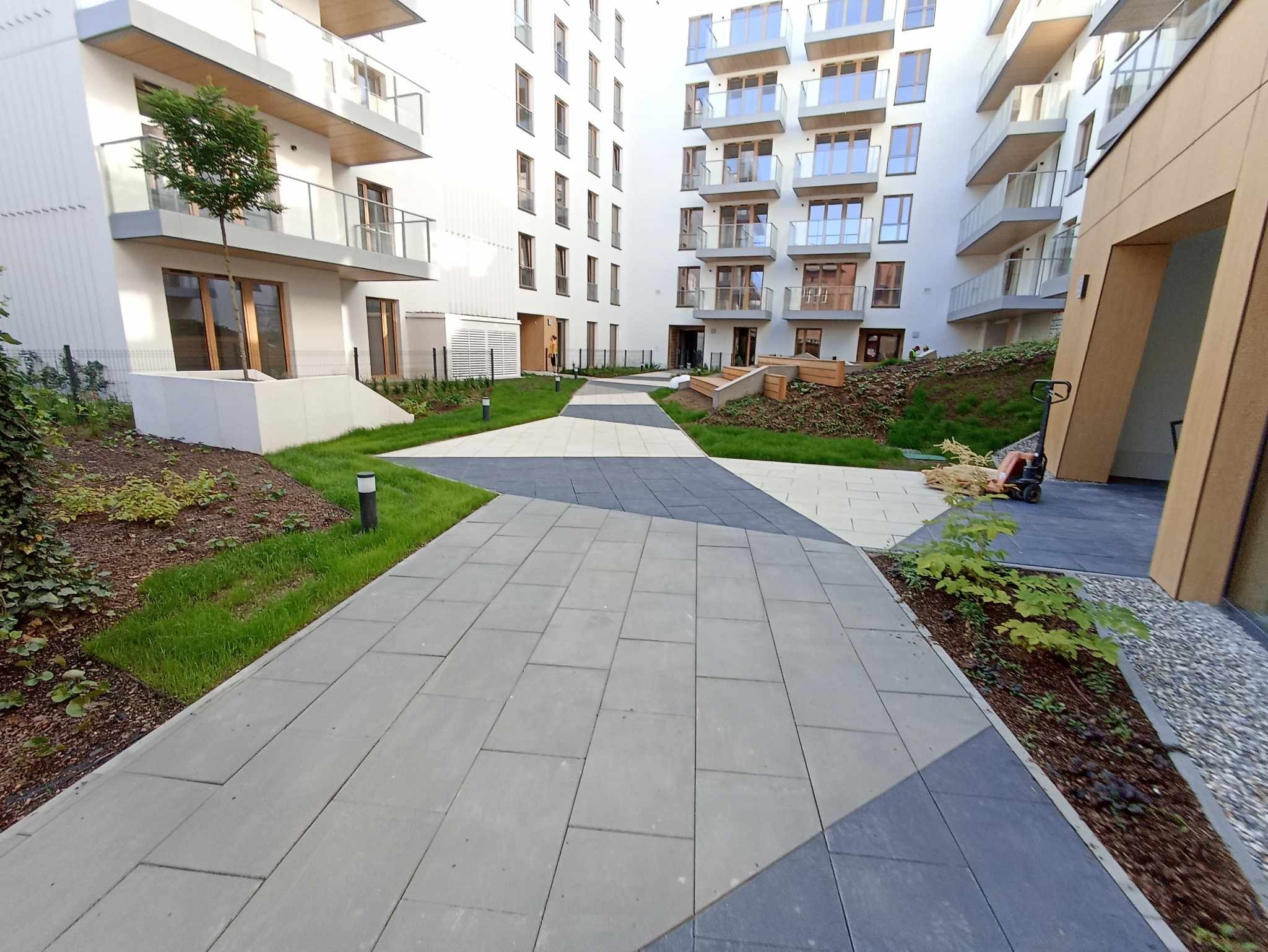 Nowoczesna ścieżka z szarych i grafitowych kostek brukowych prowadząca między budynkami apartamentowymi z zielonymi rabatami po bokach.