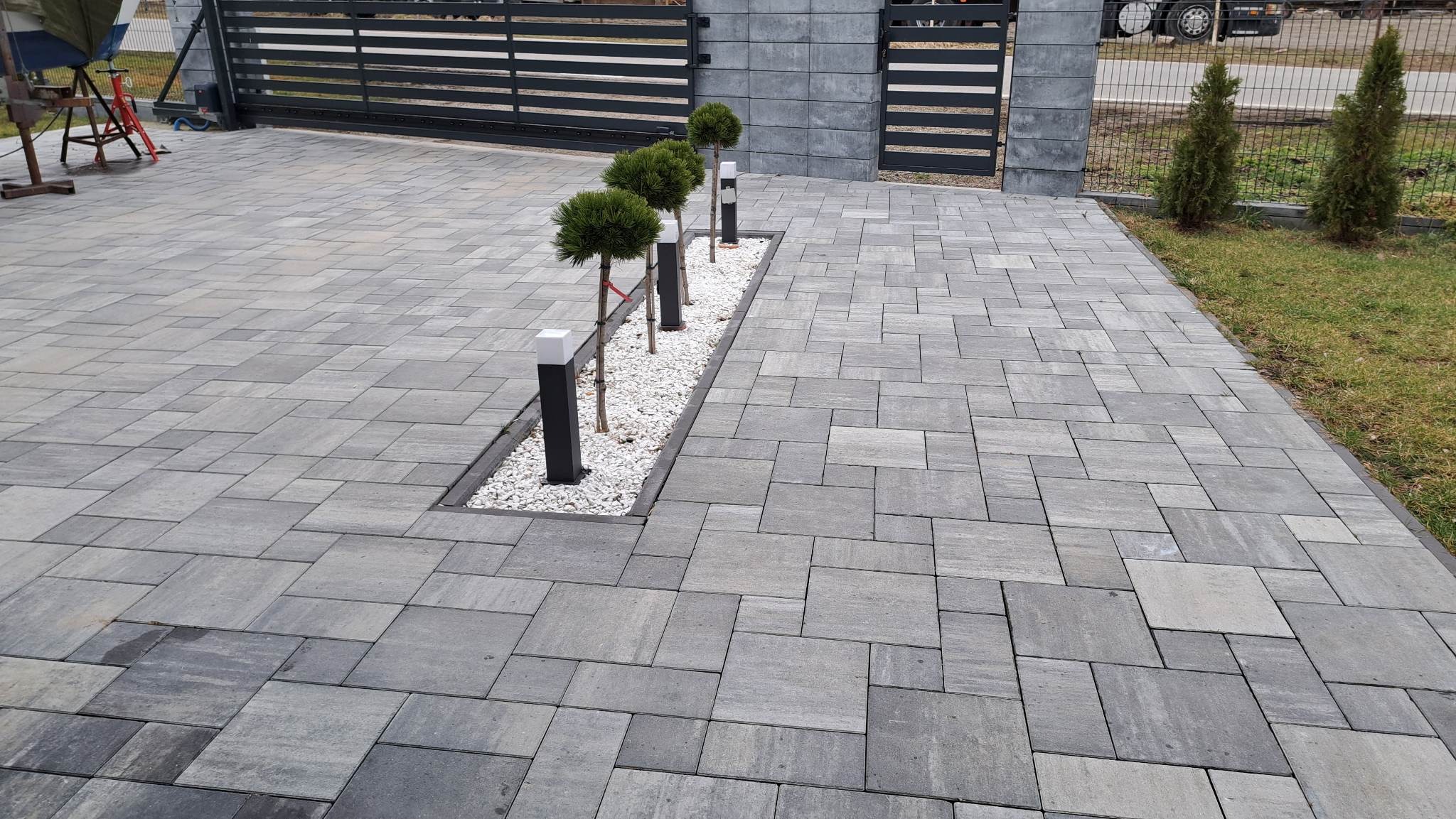 Szara kostka brukowa ułożona w geometryczny wzór na podjeździe, z wąskim pasem białego żwiru, w którym rosną trzy małe kuliste drzewka i stoją czarne lampy, brama w tle.