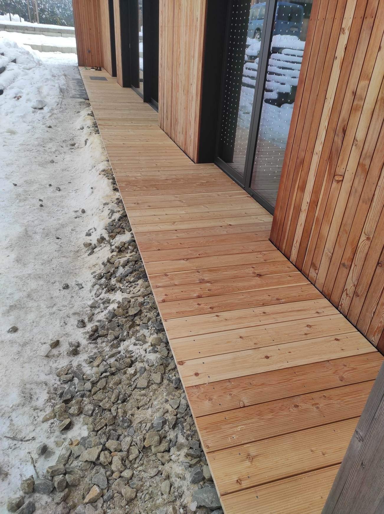Jasna, drewniana ścieżka z desek prowadząca wzdłuż ściany budynku obłożonej drewnem, obok widoczne kamienie i resztki śniegu.