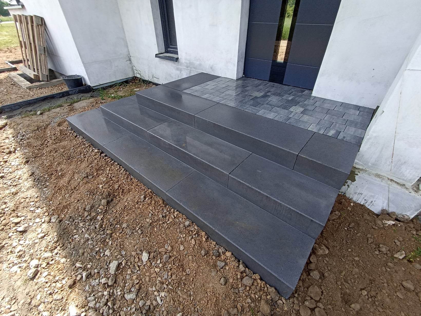 Schody wejściowe do domu, wykonane z szarych, betonowych płyt, prowadzące do drzwi wejściowych z kostki brukowej. Widoczne świeżo ułożone płyty na podłożu z ziemi i kamieni.