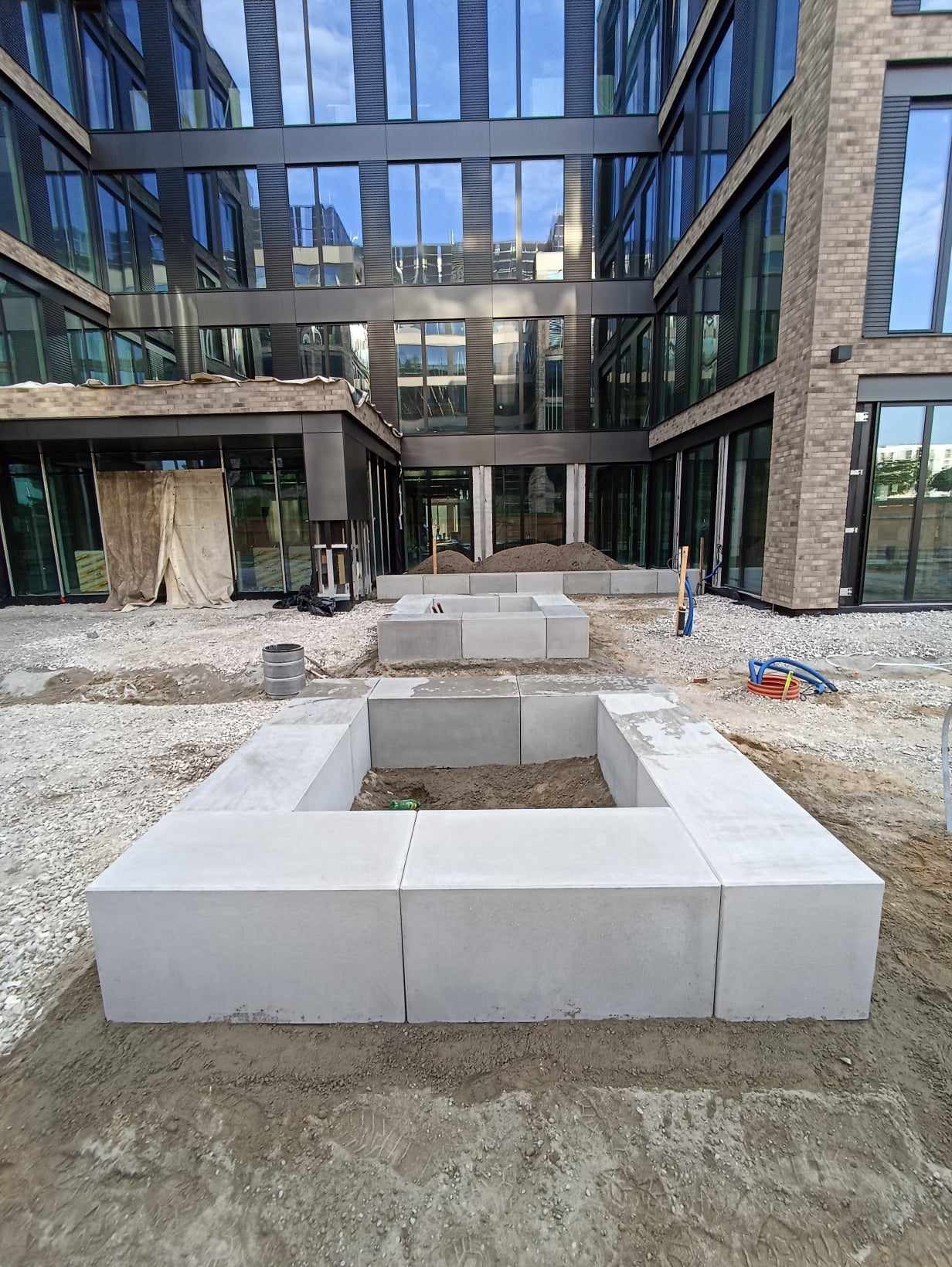 Betonowe obrzeża klombów ułożone na żwirowym podłożu przed nowoczesnym budynkiem biurowym z dużymi oknami.