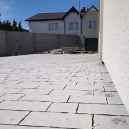 Świeżo ułożona, szara nawierzchnia z kostki brukowej na posesji, widok z niskiej perspektywy, z narzędziami i workami w tle, betonowy płot i domy w tle.