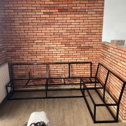 Norbert Bizewski - Metalowa konstrukcja narożnika na tle ceglanej ściany i białych płytek. Widoczny grzejnik i fragment podłogi. Surowy, industrialny styl.