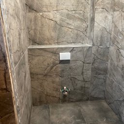 Norbert Bizewski - Wnętrze łazienki w trakcie remontu: szare płytki imitujące marmur na ścianach i podłodze, widoczne elementy instalacji i profil montażowy.