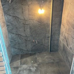 Norbert Bizewski - Wykończona łazienka w Gdyni z szarymi płytkami imitującymi marmur na ścianach i podłodze, widoczne przyłącza wodne i oświetlenie.