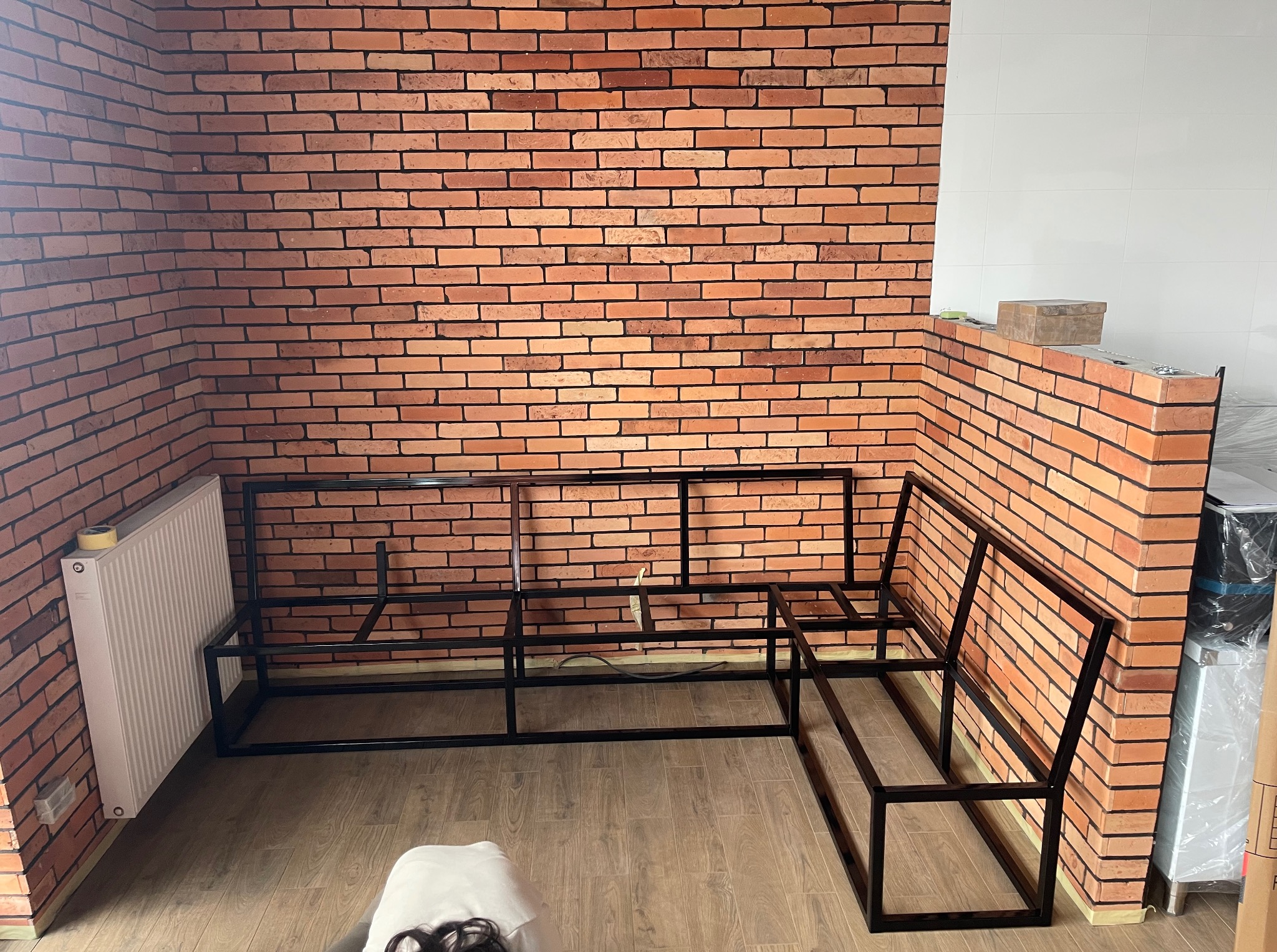 Metalowa konstrukcja narożnika na tle ceglanej ściany i białych płytek. Widoczny grzejnik i fragment podłogi. Surowy, industrialny styl.