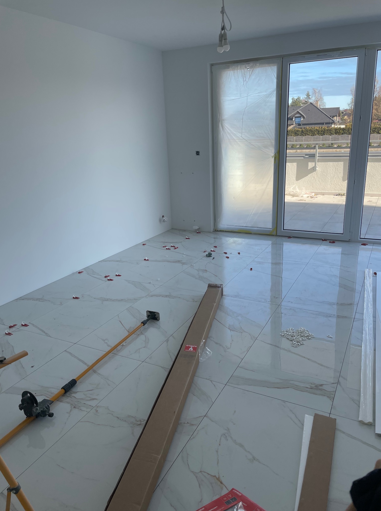 Układanie płytek podłogowych w jasnym wnętrzu. Widoczne narzędzia glazurnicze i elementy dystansowe. Okno balkonowe z widokiem na zewnątrz.