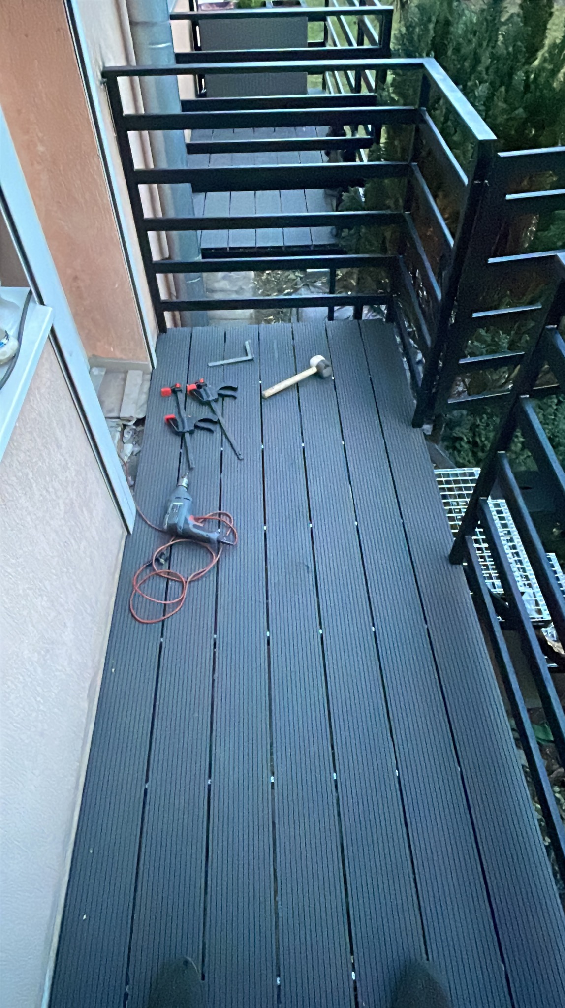 Montaż balustrady schodowej z czarnych elementów stalowych na podeście z desek kompozytowych. Widoczne narzędzia: wiertarka, młotek, ściski.