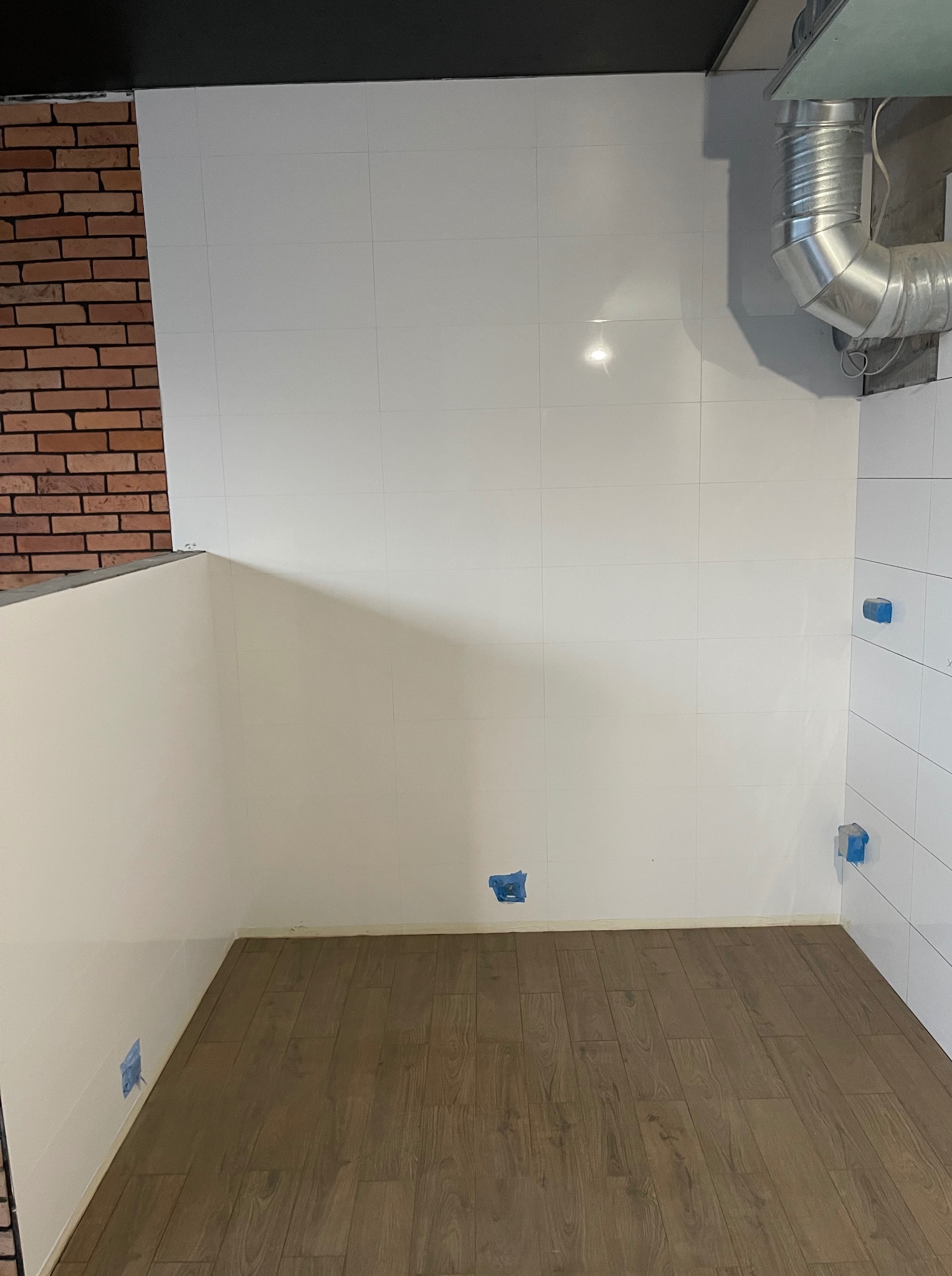 Wykończona łazienka z białymi płytkami i drewnopodobną podłogą. Widoczne fragmenty instalacji wentylacyjnej i ceglanej ściany. Niebieska taśma maskująca na ścianach.