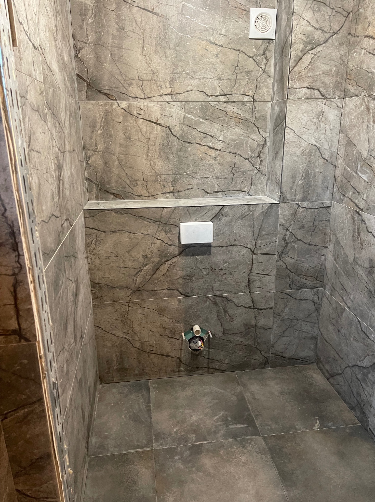 Wnętrze łazienki w trakcie remontu: szare płytki imitujące marmur na ścianach i podłodze, widoczne elementy instalacji i profil montażowy.