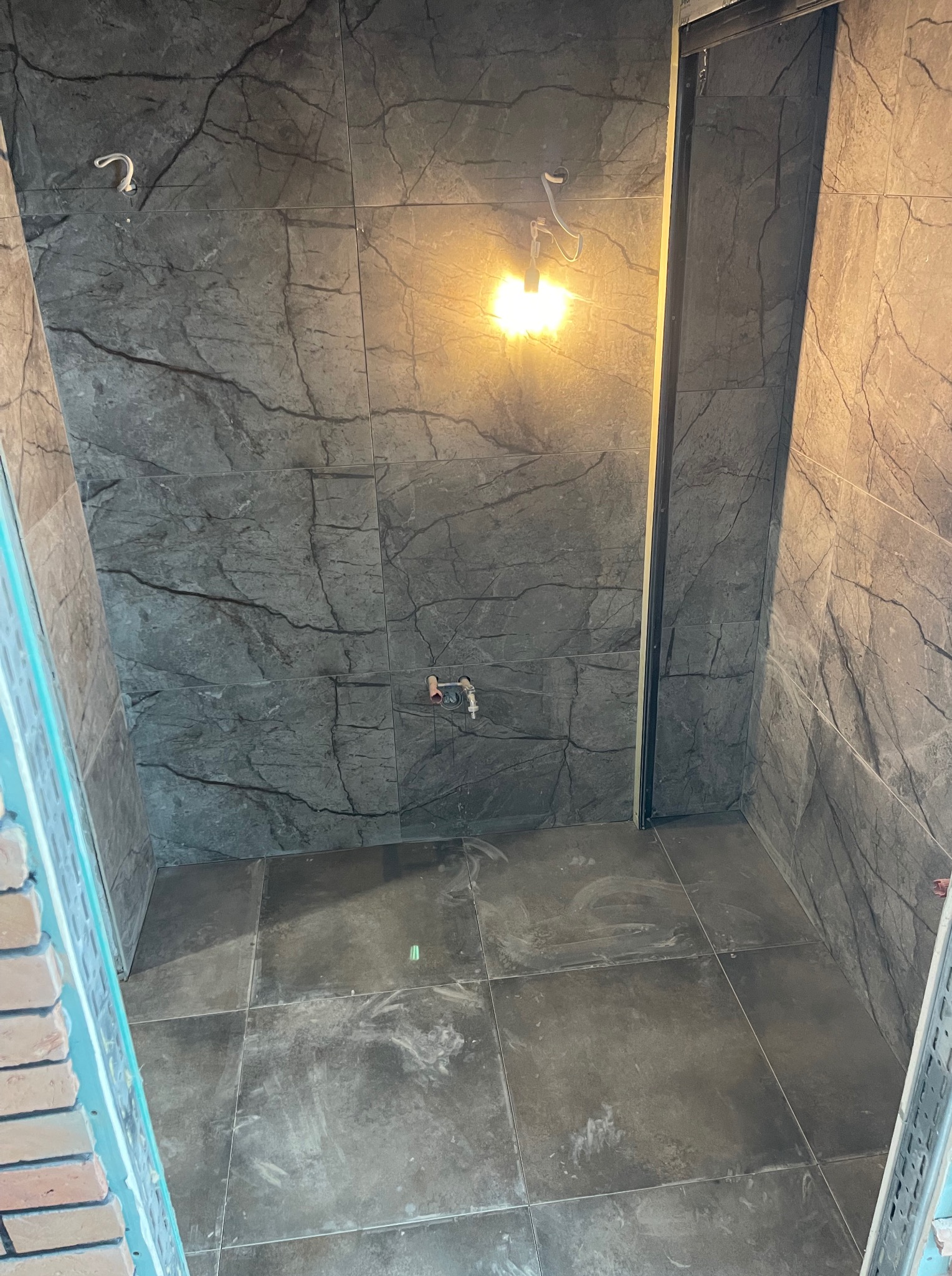 Wykończona łazienka w Gdyni z szarymi płytkami imitującymi marmur na ścianach i podłodze, widoczne przyłącza wodne i oświetlenie.