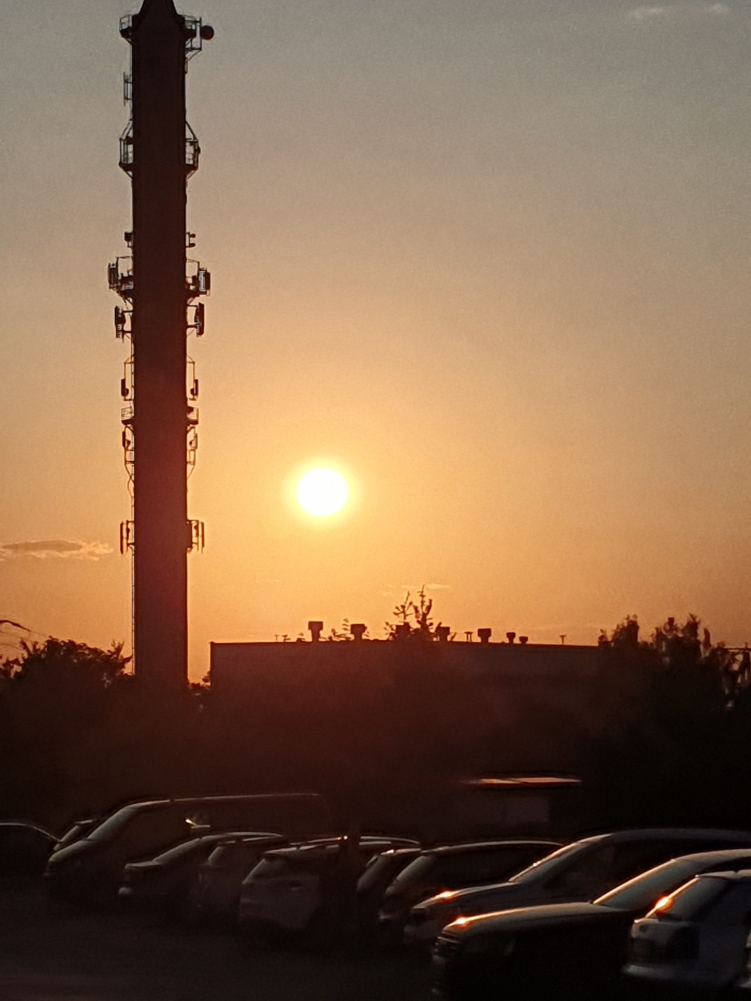 Silhouetted telecommunications tower against a sunset sky, with a building and parked cars in the foreground.