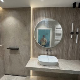 KD-BUD - Nowoczesna łazienka z umywalką nablatową, lustrem w okrągłej ramie i oświetleniem LED. Widoczny odpływ liniowy w podłodze i płytki imitujące kamień.