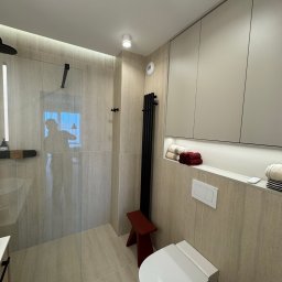 KD-BUD - Nowoczesna łazienka z kabiną prysznicową typu walk-in, beżowymi płytkami imitującymi kamień i wiszącą toaletą. Minimalistyczny design podkreślony przez czarne akcenty.