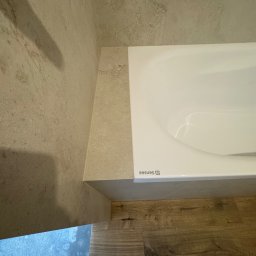 KD-BUD - Wykończona łazienka: biała wanna SENSEA z obudową z płytek imitujących kamień, drewnopodobna podłoga, ściany wyłożone płytkami w beżowym odcieniu. Ujęcie z góry.