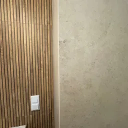 Nowoczesna łazienka: drewniane panele ścienne z przełącznikiem światła kontrastują z beżową płytką. Widoczny fragment umywalki.