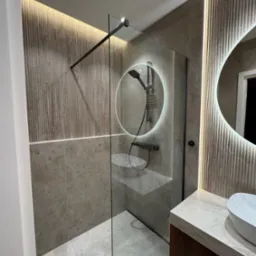 Nowoczesna łazienka z kabiną prysznicową typu walk-in, okrągłym lustrem z podświetleniem LED i umywalką nablatową. Stonowana kolorystyka, płytki imitujące kamień.
