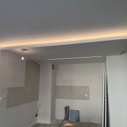 KD-BUD - Wnętrze w trakcie wykańczania: częściowo położone płytki, podwieszany sufit z oświetleniem LED, widoczne instalacje i drabina. W tle surowy beton i wisząca żarówka.