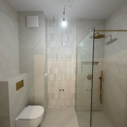 KD-BUD - Nowoczesna łazienka z kabiną prysznicową typu walk-in, wykończona płytkami imitującymi marmur i złotą armaturą. Widoczne podwieszane WC i minimalistyczny design.