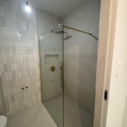 KD-BUD - Wykończona łazienka z kabiną prysznicową typu walk-in, złotymi elementami armatury i białymi, pionowo ułożonymi płytkami. Widoczne niedokończone podłączenie oświetlenia.