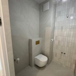 KD-BUD - Nowoczesna łazienka w odcieniach szarości z podwieszaną toaletą i płytkami imitującymi marmur. Widoczne przyłącza elektryczne i wentylacja.