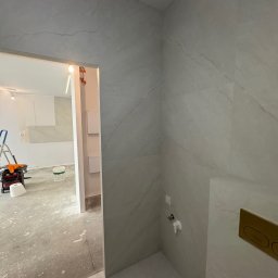 KD-BUD - Wykończona ściana łazienki z szarymi płytkami imitującymi marmur, widoczny fragment toalety i instalacja wodna. W tle pomieszczenie w trakcie remontu.