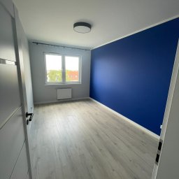 KD-BUD - Pomalowane na granatowo wnętrze pokoju z panelami podłogowymi, widok zza uchylonych drzwi, szara ściana z oknem i grzejnikiem.