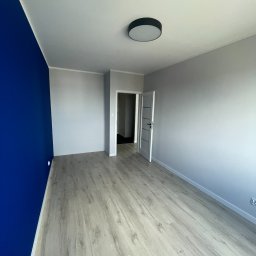 KD-BUD - Wnętrze pokoju po remoncie z jedną ścianą pomalowaną na intensywny niebieski kolor, panele podłogowe o wzorze jasnego drewna, szara ściana i białe listwy przypodłogowe, widoczne drzwi do kolejnego...