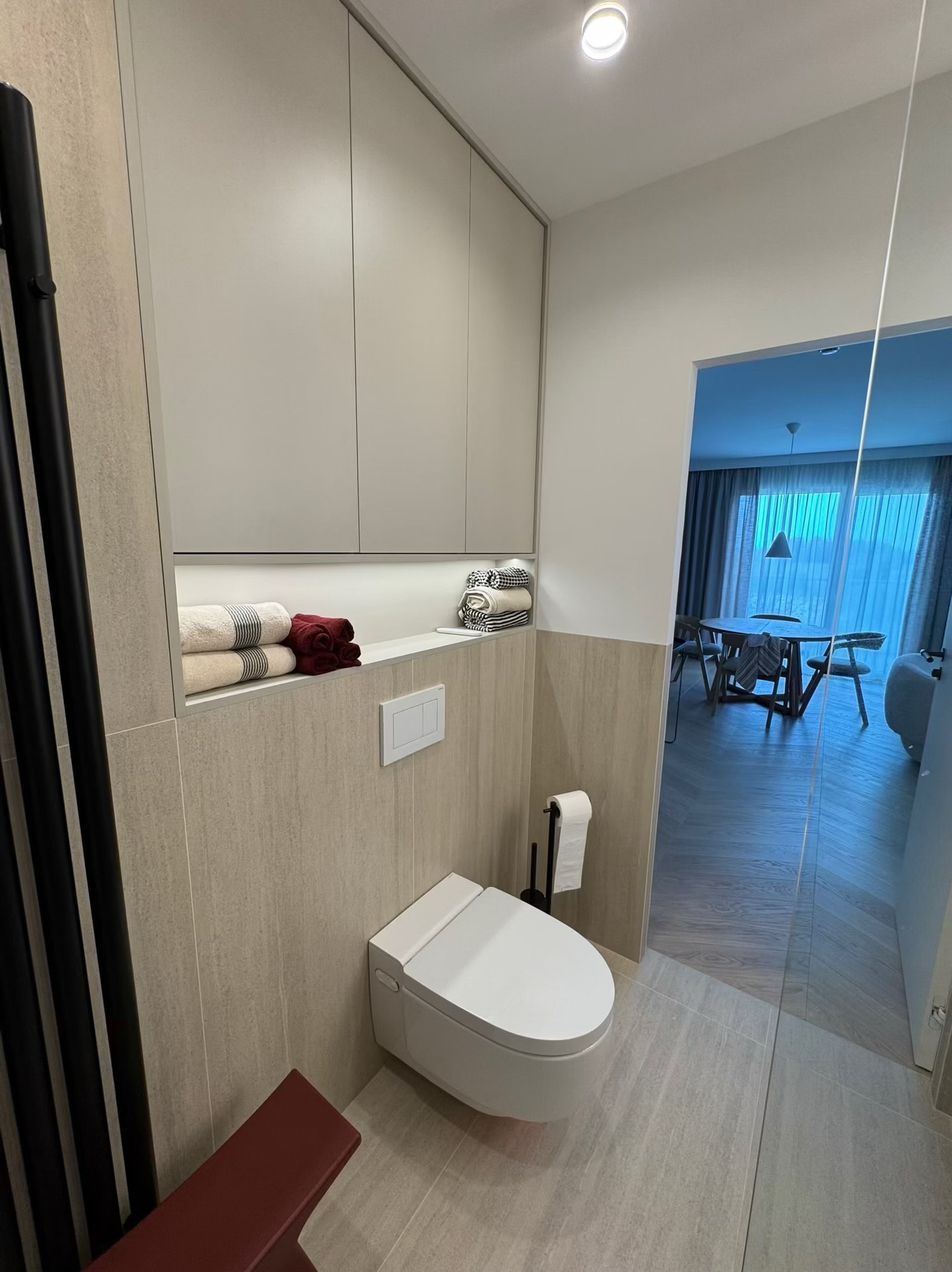 Nowoczesna łazienka z podwieszaną toaletą, beżowymi płytkami i minimalistyczną zabudową meblową. W tle widoczny salon z jadalnią, oddzielony szklaną ścianą.