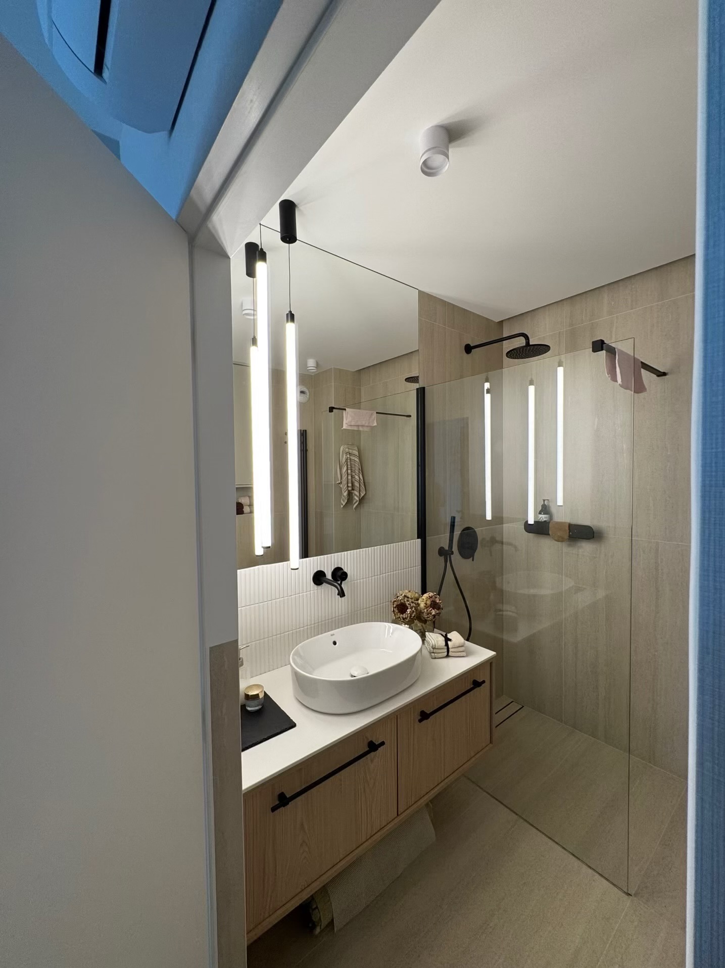 Nowoczesna łazienka z owalną umywalką, drewnianą szafką i kabiną prysznicową oddzieloną szkłem. Minimalistyczne oświetlenie LED i czarne akcenty armatury.