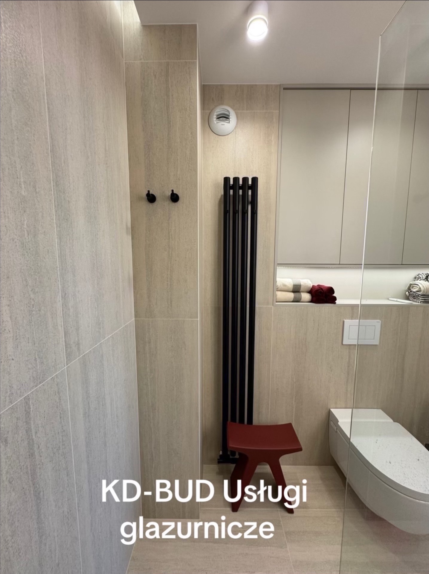Nowoczesna łazienka z beżowymi płytkami, czarnym grzejnikiem drabinkowym i czerwonym stołkiem. Widoczny fragment toalety i szklana kabina prysznicowa.