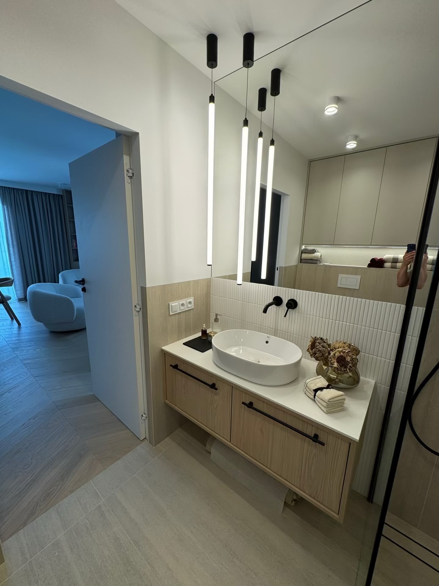 Nowoczesna łazienka z owalną umywalką nablatową, czarną armaturą i wiszącymi lampami tubami. W tle widoczne lustro i fragment salonu.