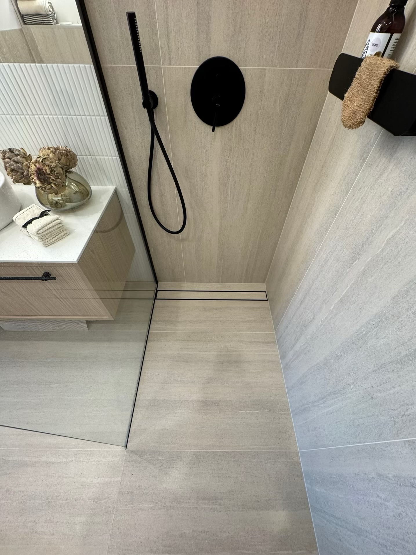 Nowoczesna kabina prysznicowa z czarną armaturą, odpływem liniowym i beżowymi płytkami imitującymi kamień. Umywalka z drewnianą szafką i dekoracjami.