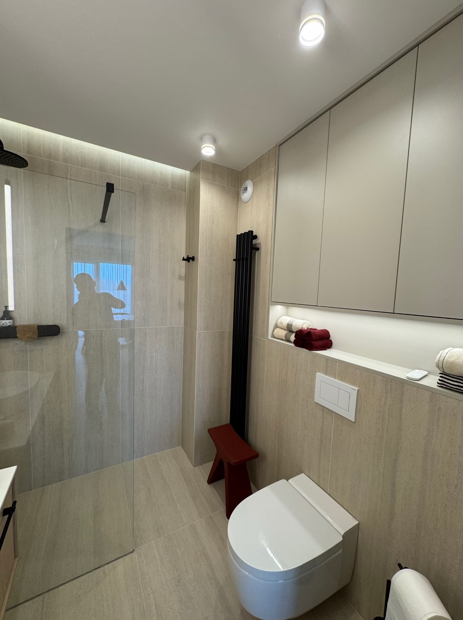 Nowoczesna łazienka z kabiną prysznicową typu walk-in, beżowymi płytkami imitującymi kamień i wiszącą toaletą. Minimalistyczny design podkreślony przez czarne akcenty.