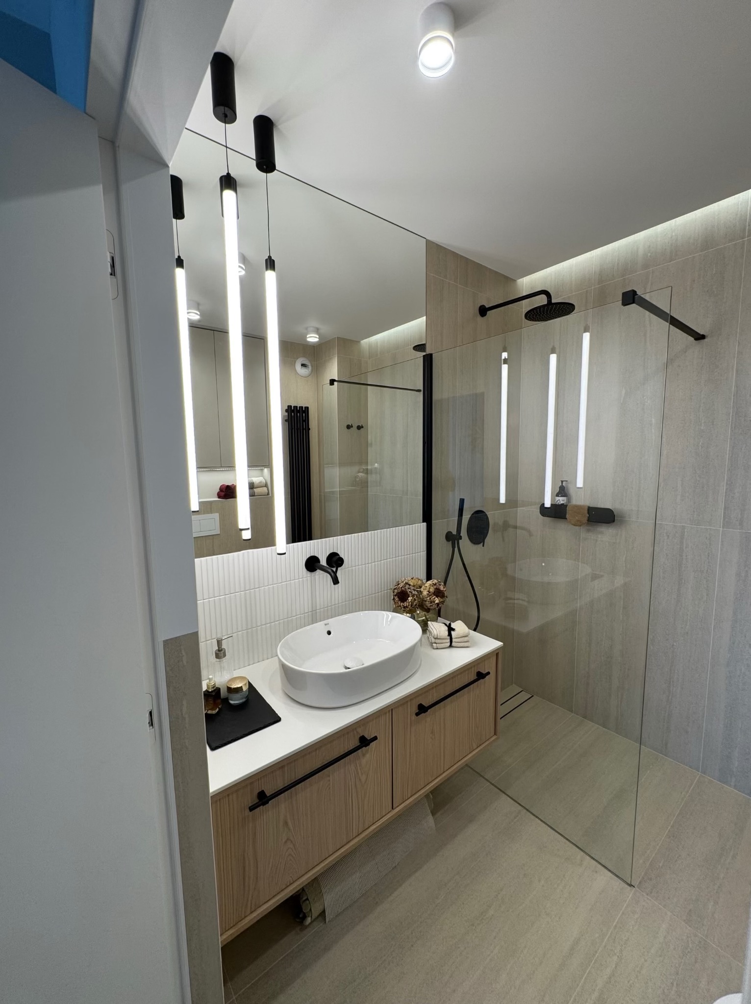 Nowoczesna łazienka: owalna umywalka na szafce z drewnianym frontem, szklana kabina prysznicowa i minimalistyczne, wiszące lampy nad lustrem. Jasna tonacja.