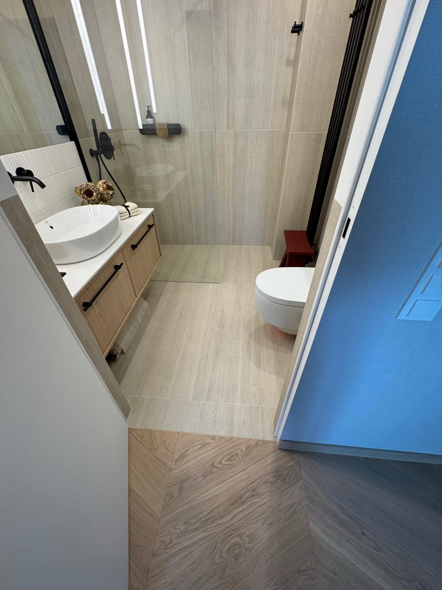Nowoczesna łazienka z drewnianą szafką pod umywalką, beżowymi płytkami i toaletą. Uwagę przykuwa parkiet ułożony w jodełkę przy wejściu.