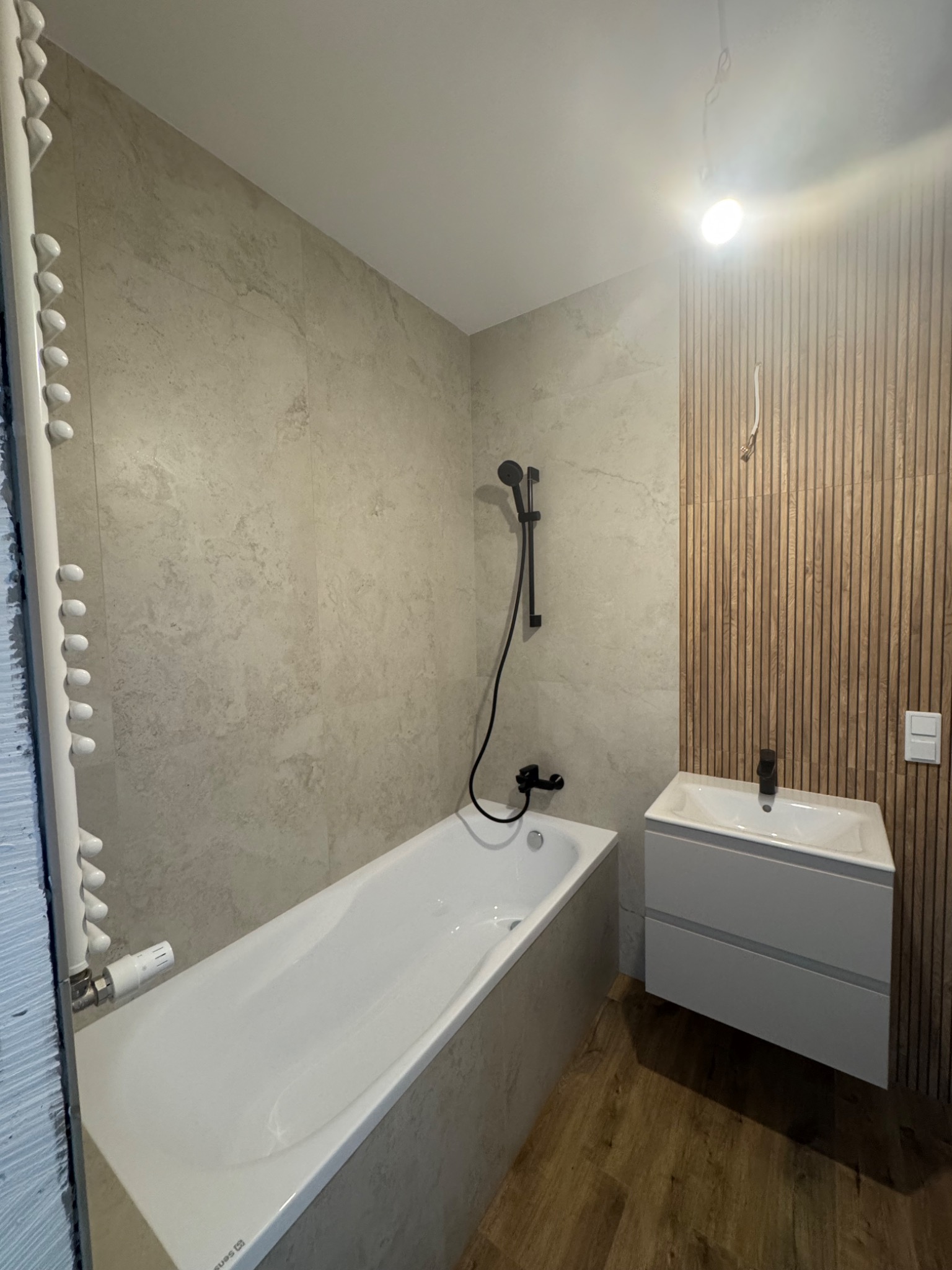 Nowoczesna łazienka z wanną, umywalką na szafce i grzejnikiem drabinkowym. Ściany wyłożone płytkami imitującymi kamień, drewniana okładzina ścienna.