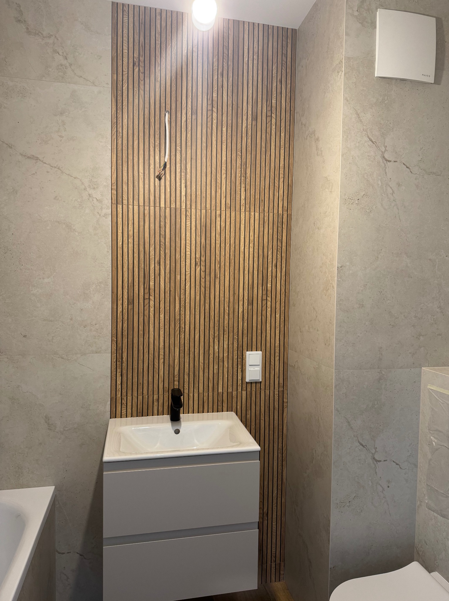 Nowoczesna łazienka z drewnianą ścianą z lameli, szarą umywalką i płytkami imitującymi kamień. Widoczny fragment wanny i toalety. Minimalistyczny design.