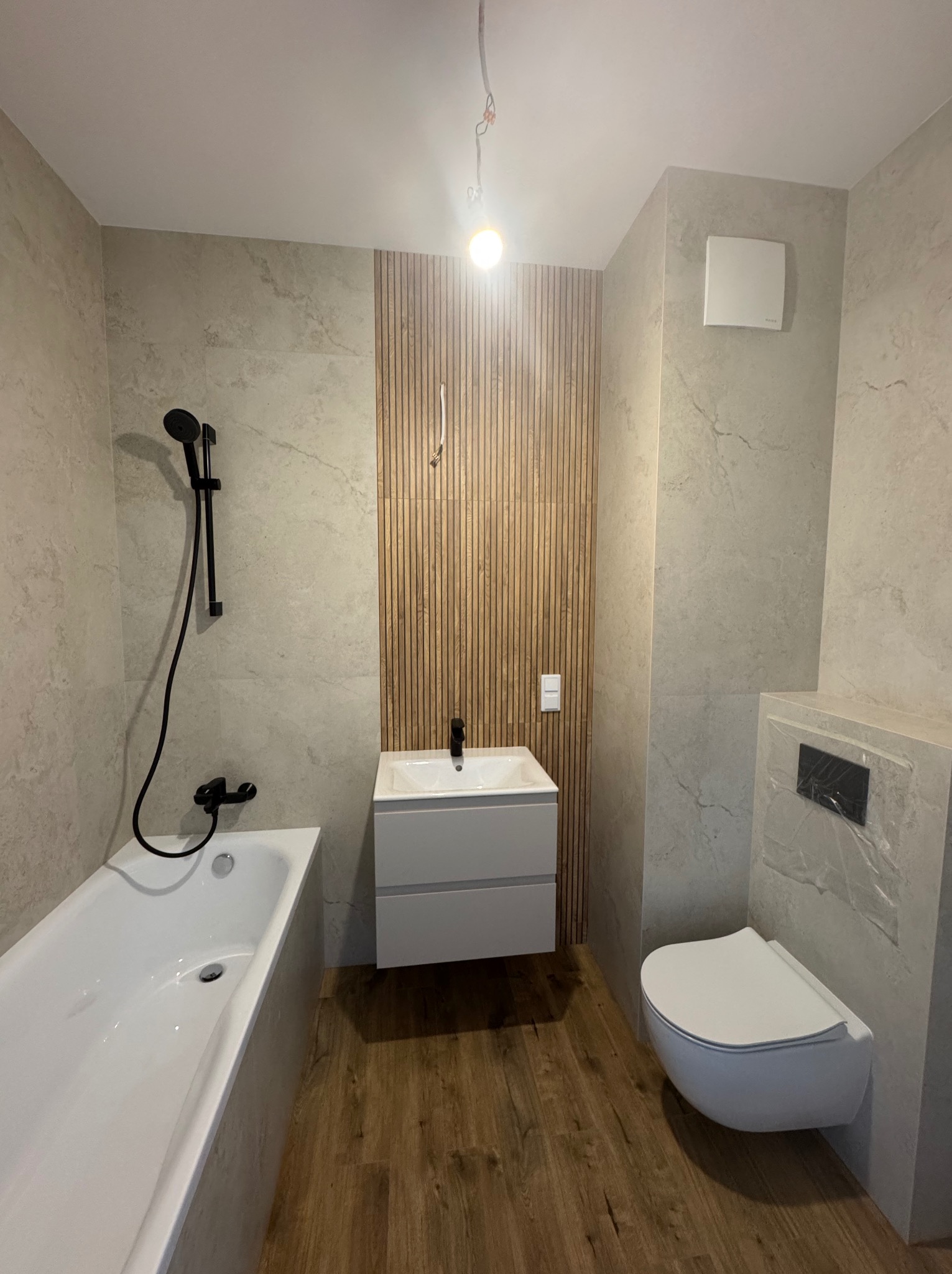 Nowoczesna łazienka z drewnianymi lamelami za umywalką, szarą armaturą i płytkami imitującymi kamień. Umywalka z szafką, wisząca toaleta i wanna.