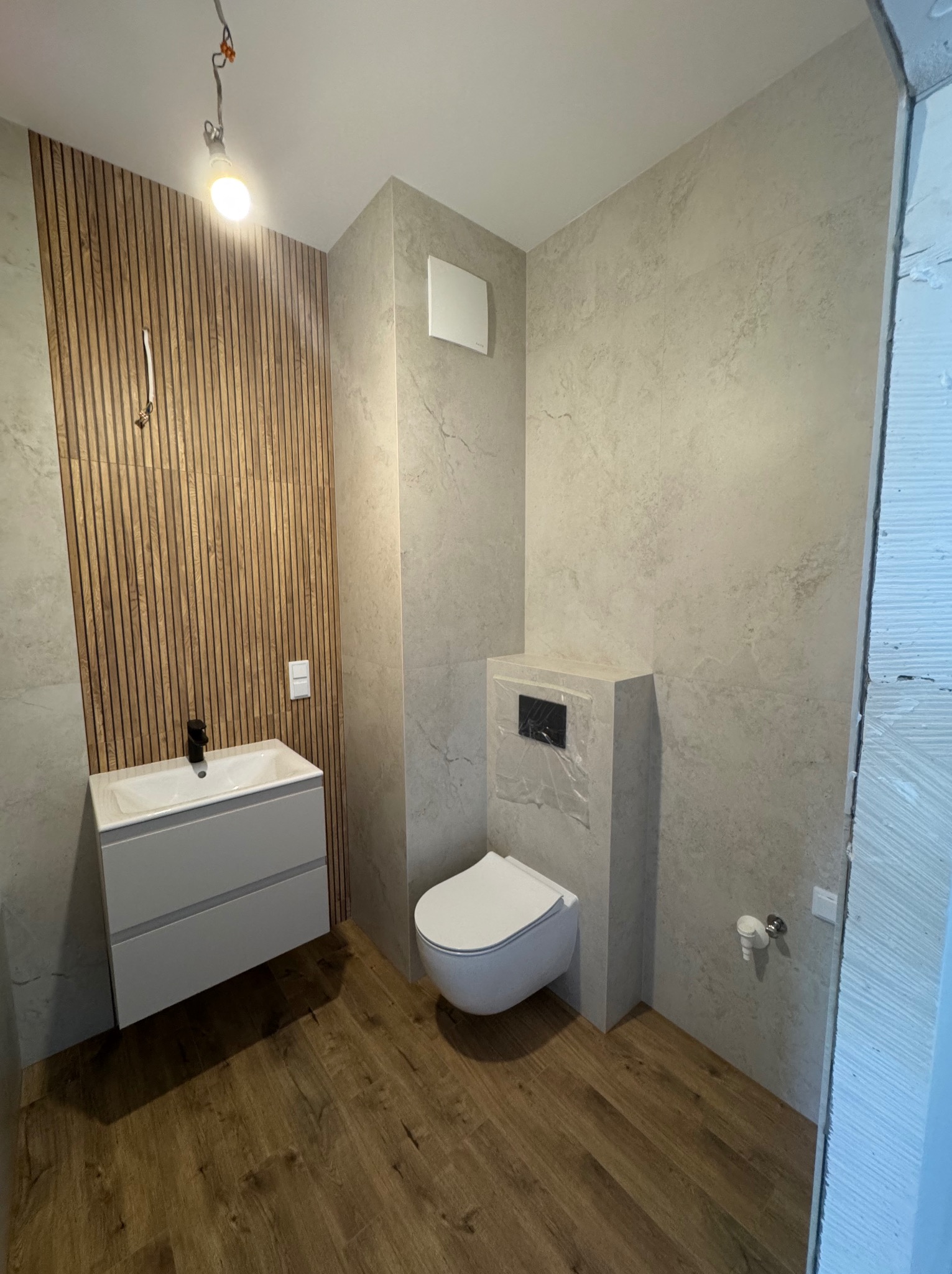 Nowoczesna łazienka z drewnianą ścianą z lameli, szarą armaturą i podwieszaną toaletą. Na suficie widoczne kable. Podłoga wyłożona panelami imitującymi drewno.