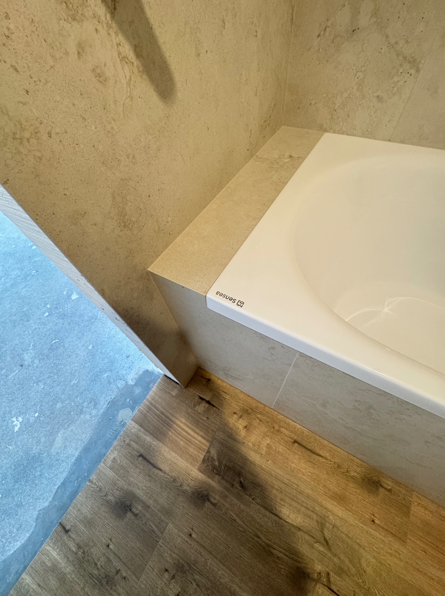 Biała wanna Sensaa, obudowana beżowymi płytkami, z widoczną podłogą w panelach drewnopodobnych i fragmentem ściany z beżowych płytek. Ukończona łazienka.