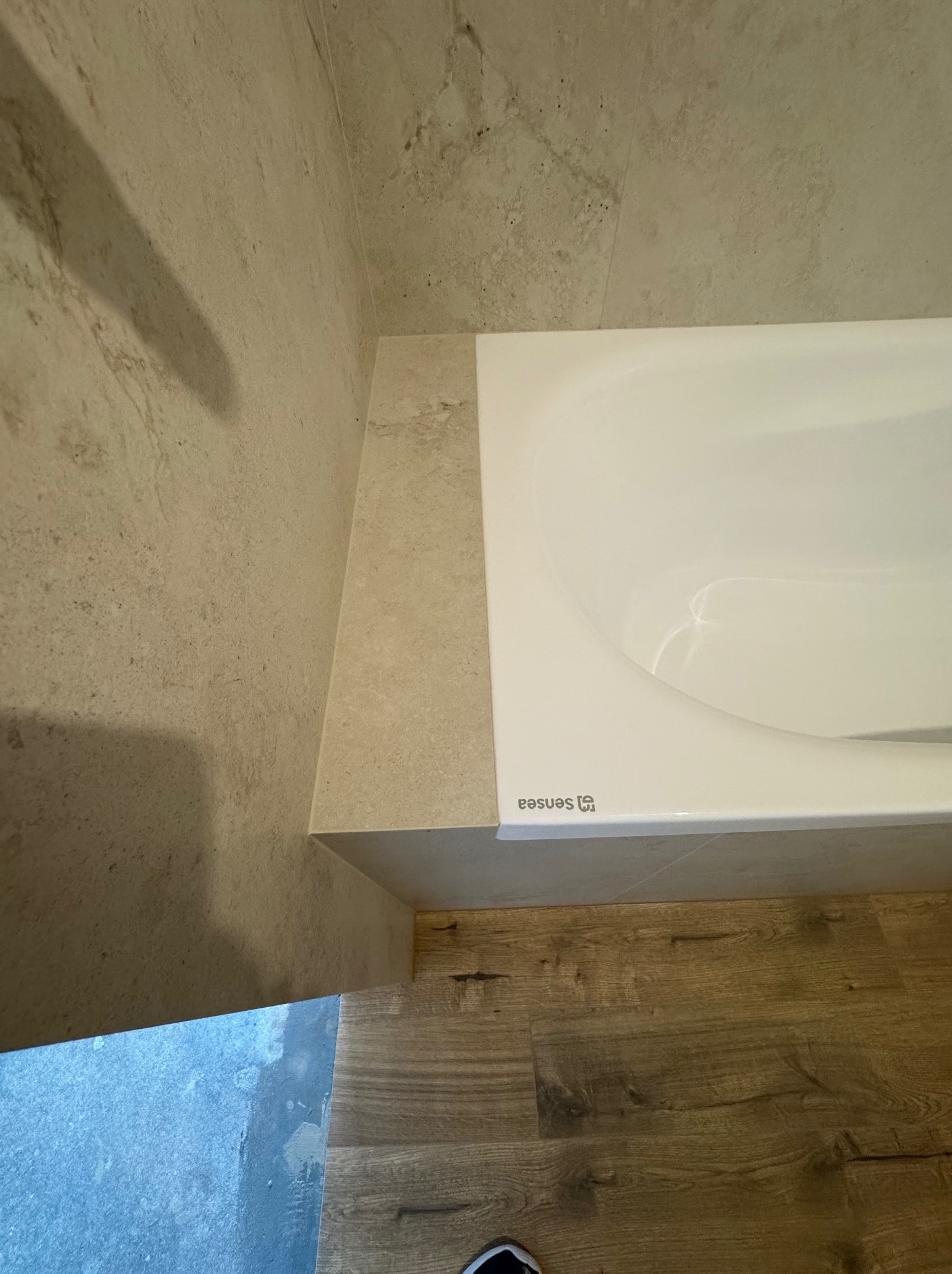 Wykończona łazienka: biała wanna SENSEA z obudową z płytek imitujących kamień, drewnopodobna podłoga, ściany wyłożone płytkami w beżowym odcieniu. Ujęcie z góry.