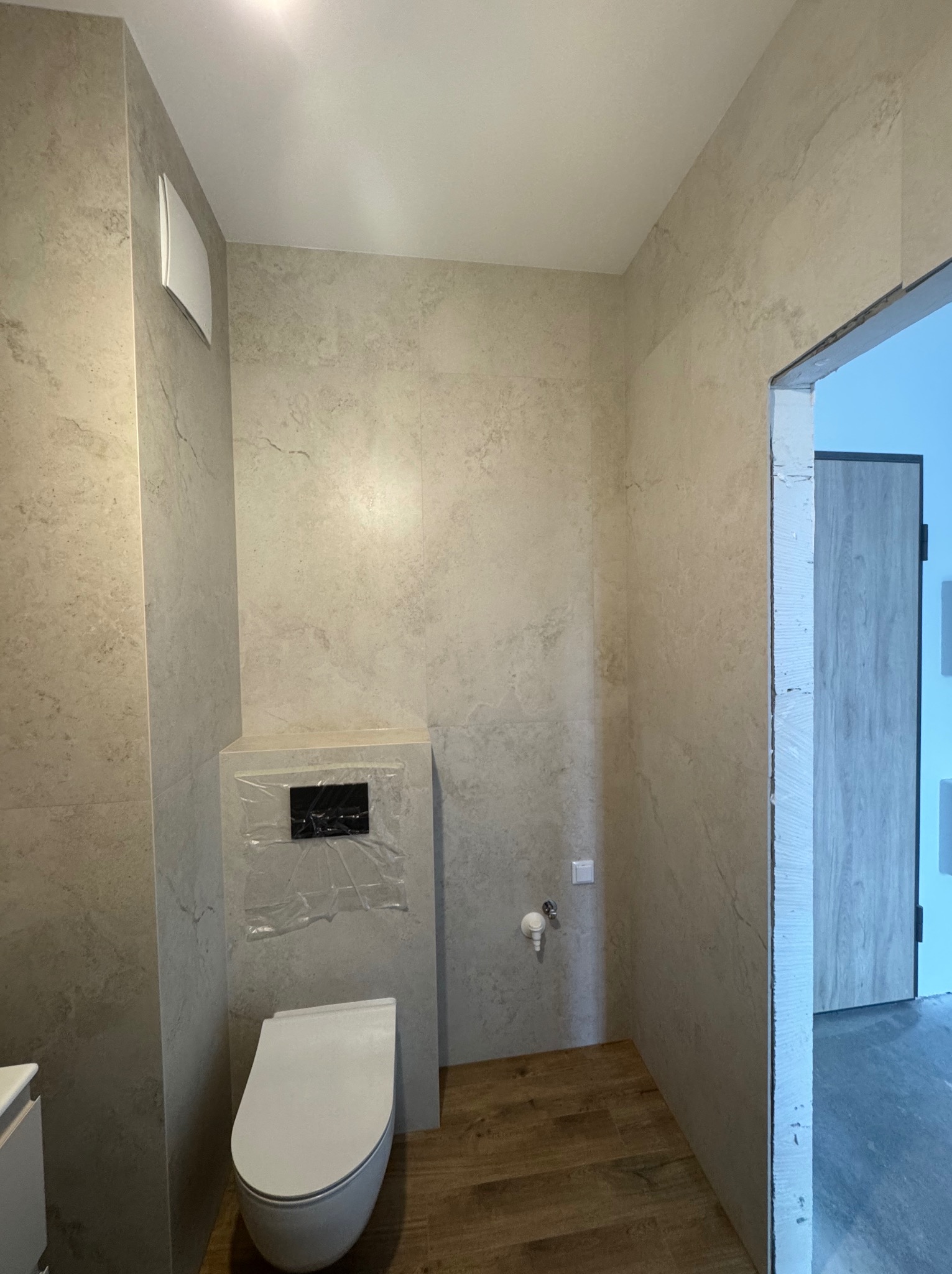 Wykończona łazienka z beżowymi płytkami imitującymi kamień, podwieszaną toaletą i drewnopodobną podłogą. Widoczny fragment drzwi i wentylacja.