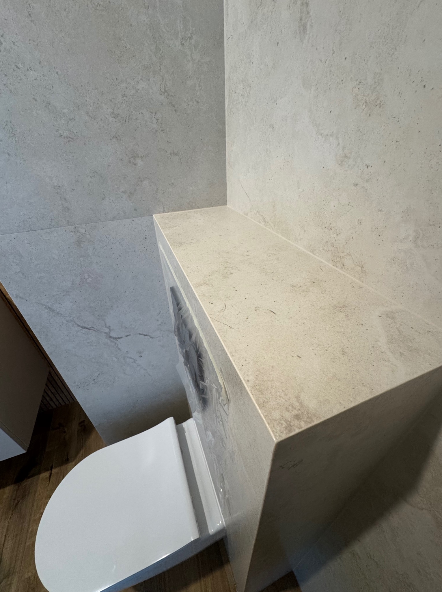 Nowoczesna toaleta podwieszana z zabudową z płytek imitujących kamień w odcieniach szarości i beżu. Widoczna deska sedesowa i fragment drewnianej podłogi.