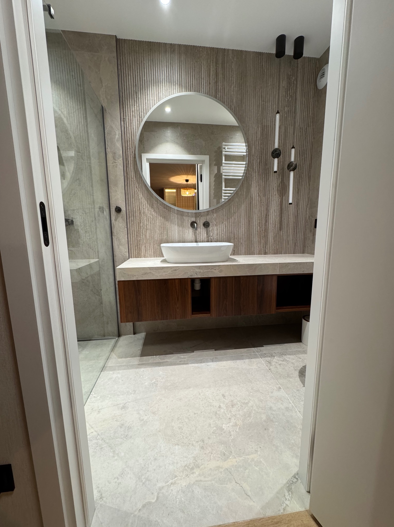Nowoczesna łazienka z okrągłym lustrem, umywalką nablatową i drewnianą szafką pod umywalką. Ściany wykończone płytkami imitującymi drewno. Widok zza uchylonych drzwi.