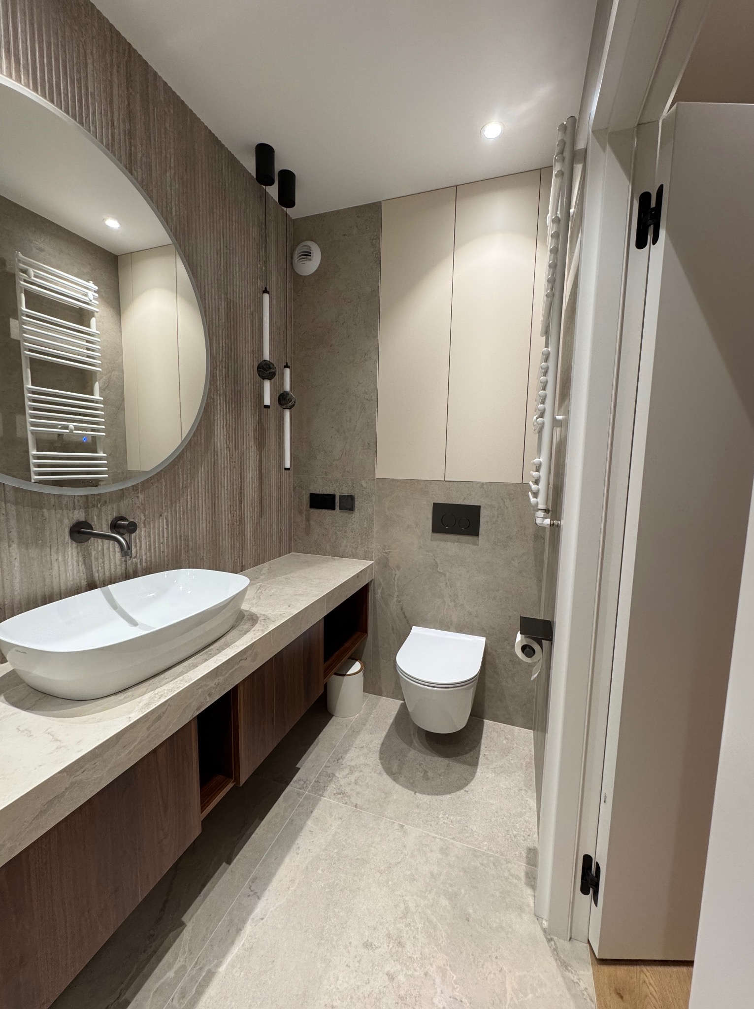 Nowoczesna łazienka z owalną umywalką nablatową, okrągłym lustrem i grzejnikiem drabinkowym. Beżowo-szara kolorystyka, minimalistyczne oświetlenie.