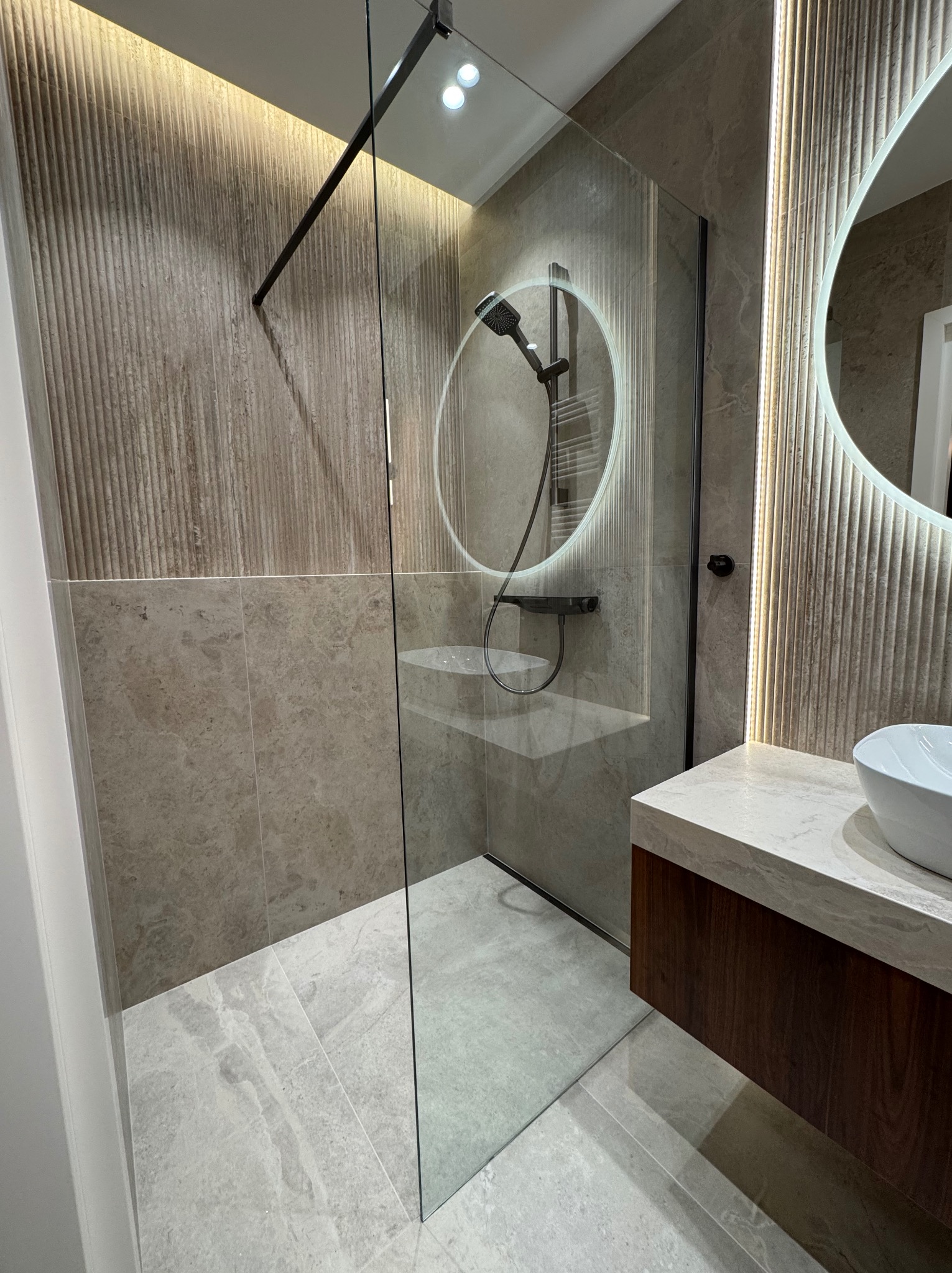 Nowoczesna łazienka z kabiną prysznicową typu walk-in, podświetlanym lustrem i umywalką nablatową. Widoczne płytki w odcieniach beżu i szarości oraz drewniana szafka pod umywalką.