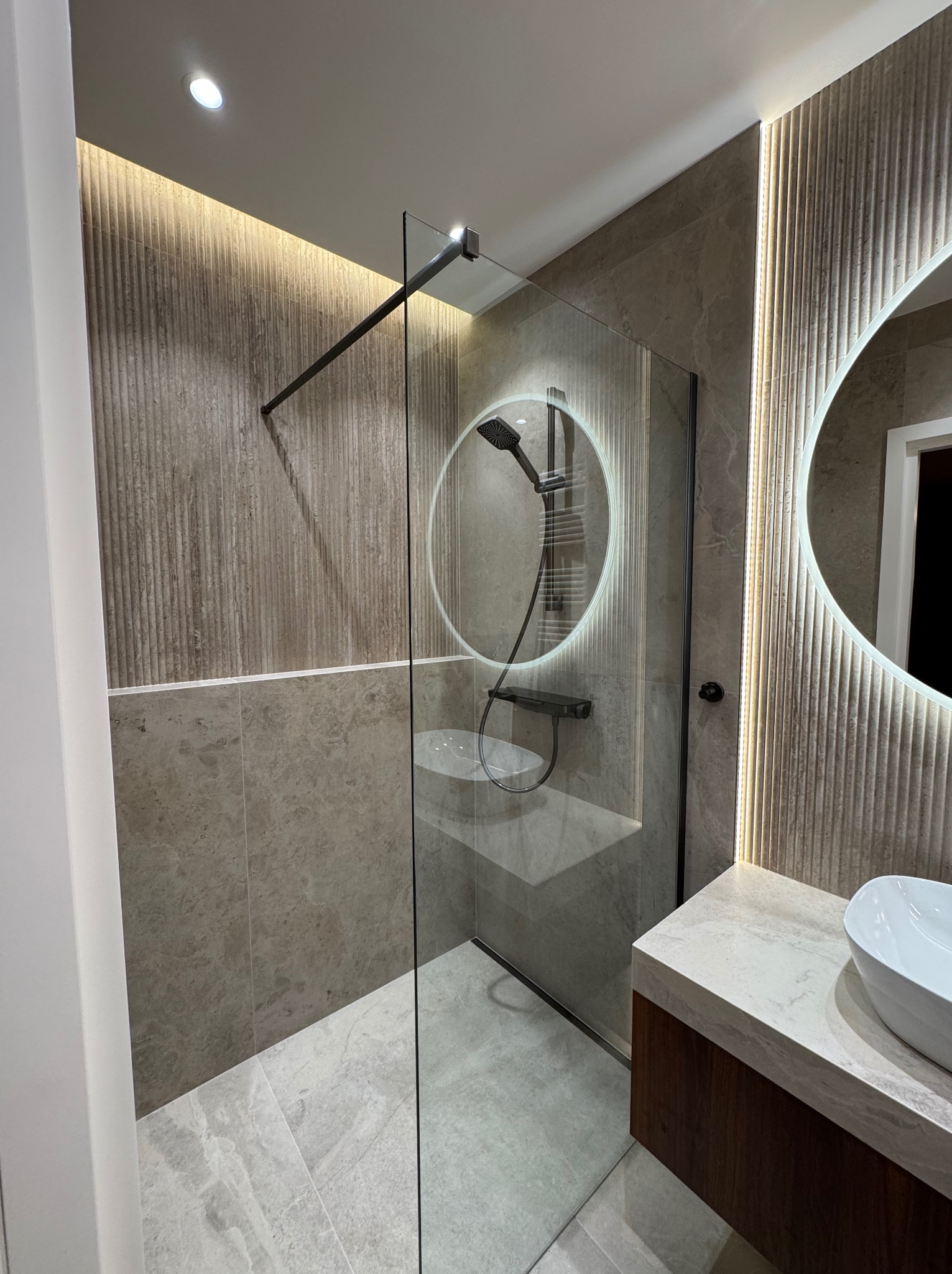 Nowoczesna łazienka z kabiną prysznicową typu walk-in, okrągłym lustrem z podświetleniem LED i umywalką nablatową. Stonowana kolorystyka, płytki imitujące kamień.